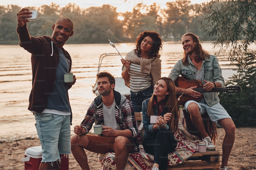Gruppe junger Leute in Freizeitkleidung, die lächeln und Selfies machen, während sie eine Strandparty in der Nähe des Sees genießen