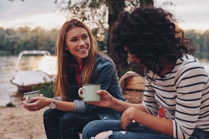 Two young beautiful women in casual wear smiling and talking while enjoying camping near the lake