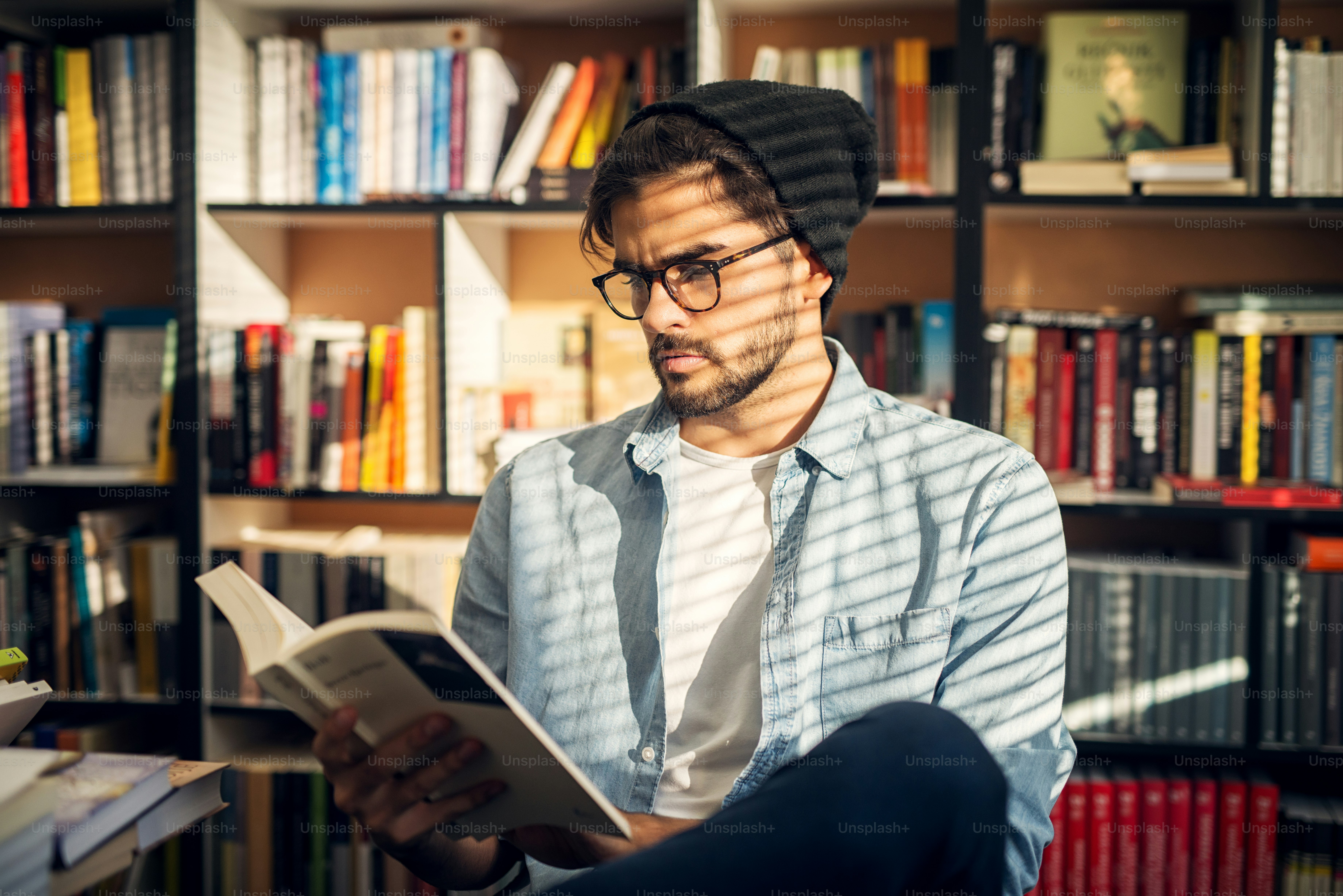 Netter Hipster-Junge, der auf dem Boden einer Bibliothek sitzt und mit neugierigem Gesicht durch einige Bücher blättert.