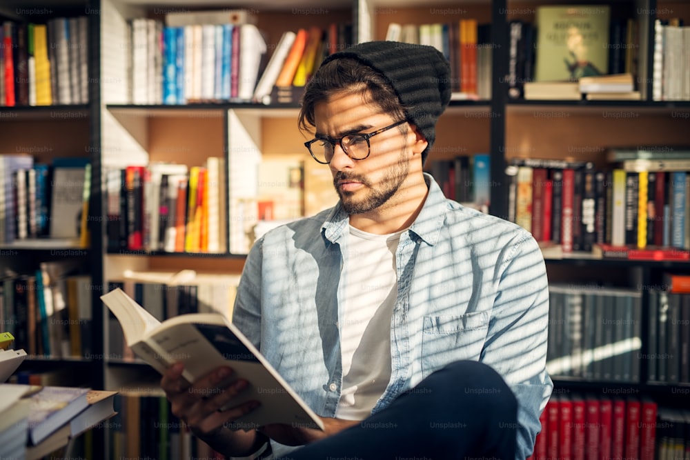 Menino hipster bonito sentado no chão de uma biblioteca e olhando através de alguns livros com cara curiosa.
