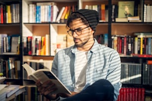 Netter Hipster-Junge, der auf dem Boden einer Bibliothek sitzt und mit neugierigem Gesicht durch einige Bücher blättert.