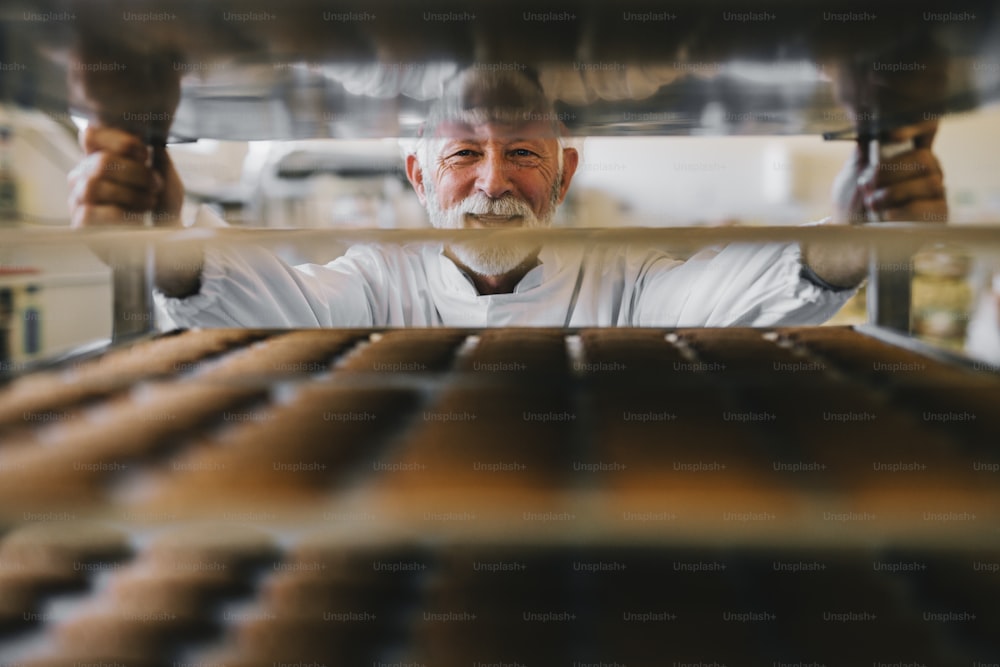 Imagen de un panadero profesional maduro con uniforme de trabajo blanco mirando a la cámara. De pie frente a los estantes llenos de galletas recién horneadas.