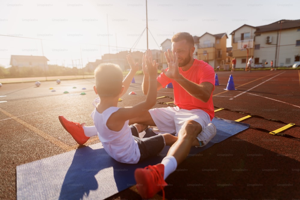 Proud young father motivating his little son to train with him. Sitting outside on training field and doing crunches.
