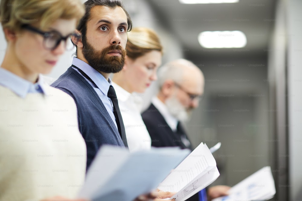 Young bearded man with documents thinking of something among his colleagues