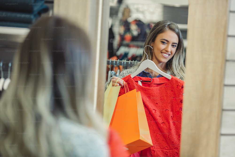 Happy woman choosing clothes and looking to mirror in mall or clothing store