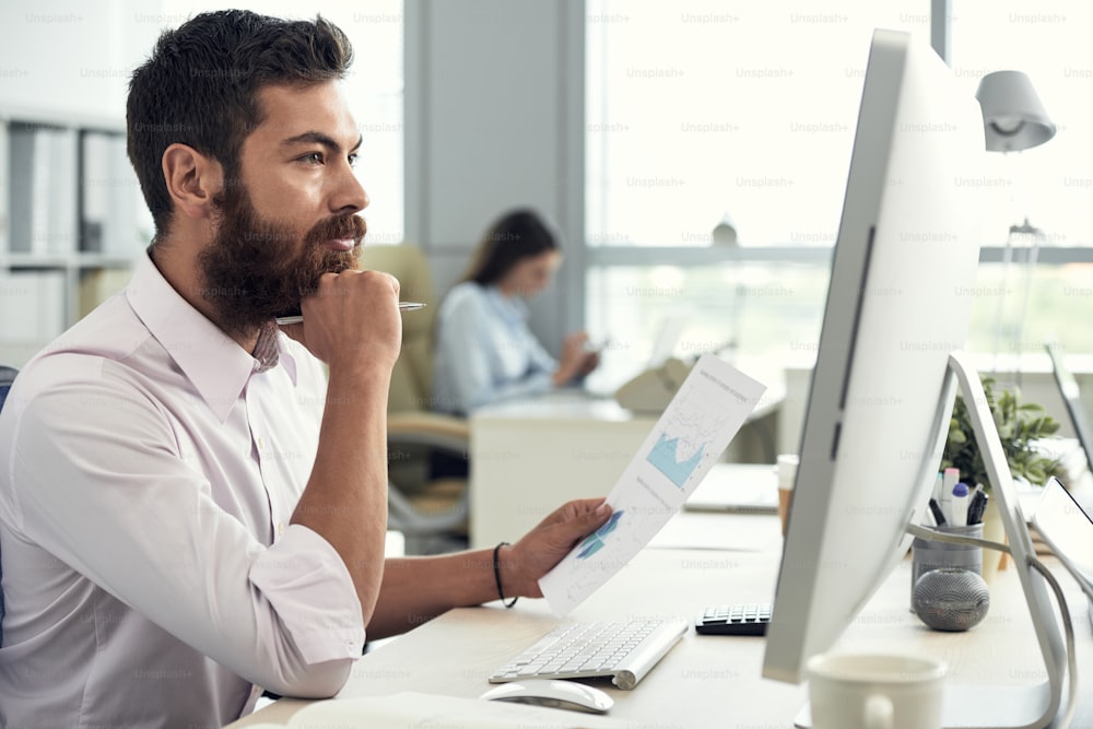 Handsome pensive entrepreneur working with documents and analyzing data on computer screen