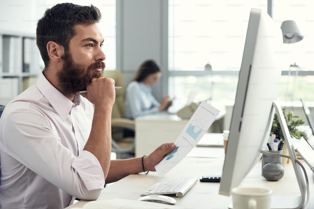 Gutaussehender nachdenklicher Unternehmer, der mit Dokumenten arbeitet und Daten auf dem Computerbildschirm analysiert