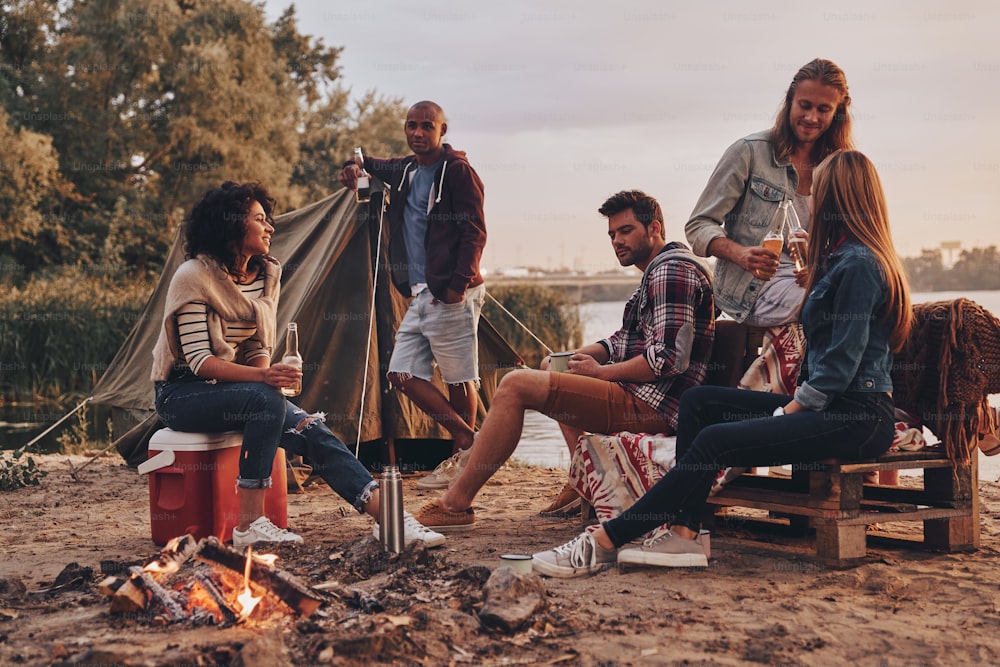 Grupo de jóvenes en ropa casual sonriendo y bebiendo cerveza mientras acampan cerca del lago