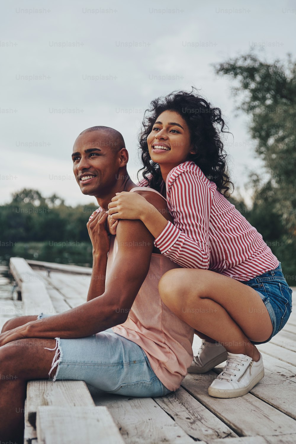 Feliz pareja joven abrazada y sonriendo mientras está sentada en el muelle cerca del lago