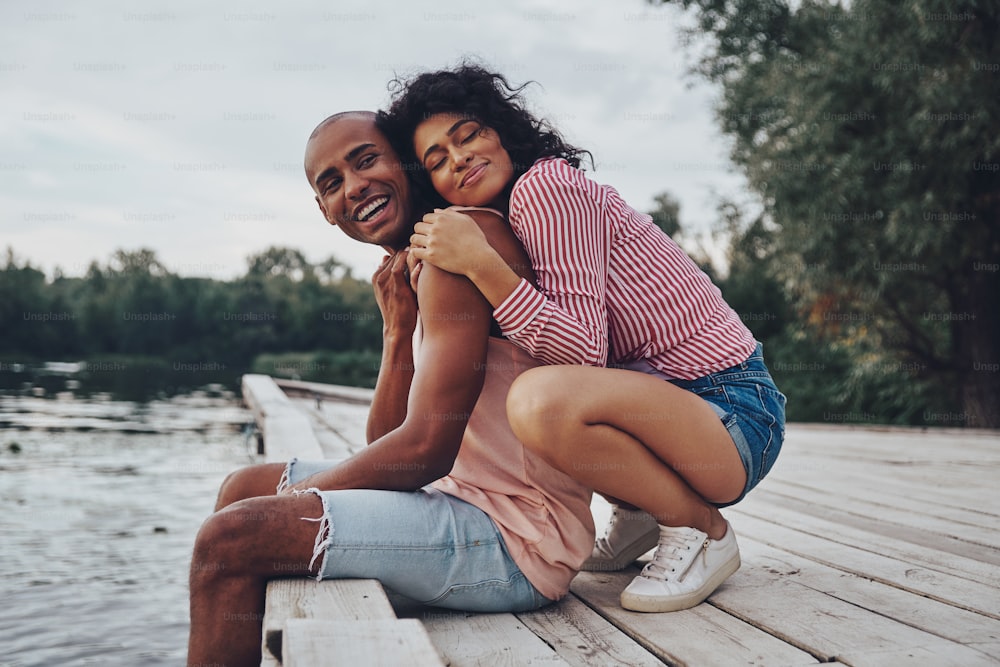 Feliz pareja joven abrazada y sonriendo mientras está sentada en el muelle cerca del lago