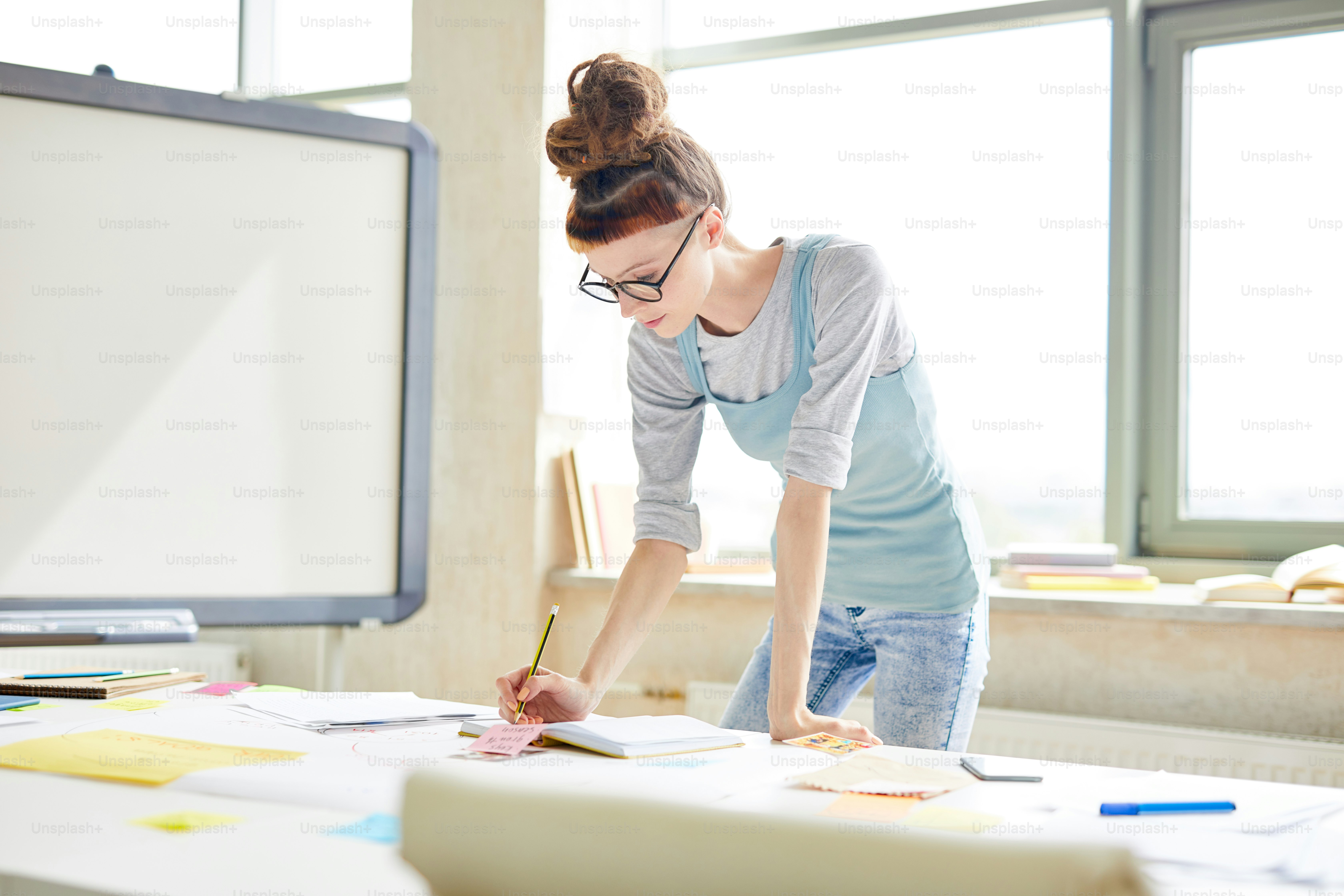 カジュアルな服と眼鏡をかけた恐怖を持つ深刻な流行に敏感なマネージャーは、現代のオフィスで新しいプロジェクトを計画しながら、主催者にメモを取ります