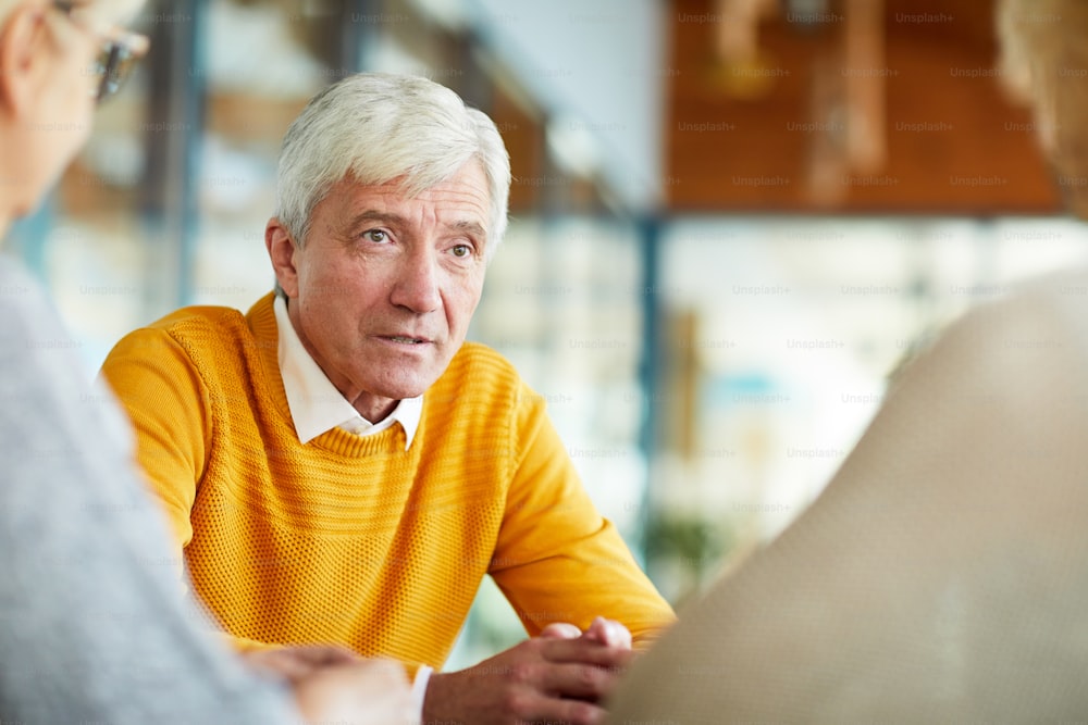 Serious confident senior man with gray hair wearing yellow sweater talking to colleagues while they discussing business in cafe