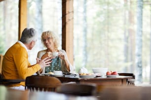 Positive beautiful mature couple in casual clothing sitting at table and drinking coffee at veranda while chatting in tranquil place