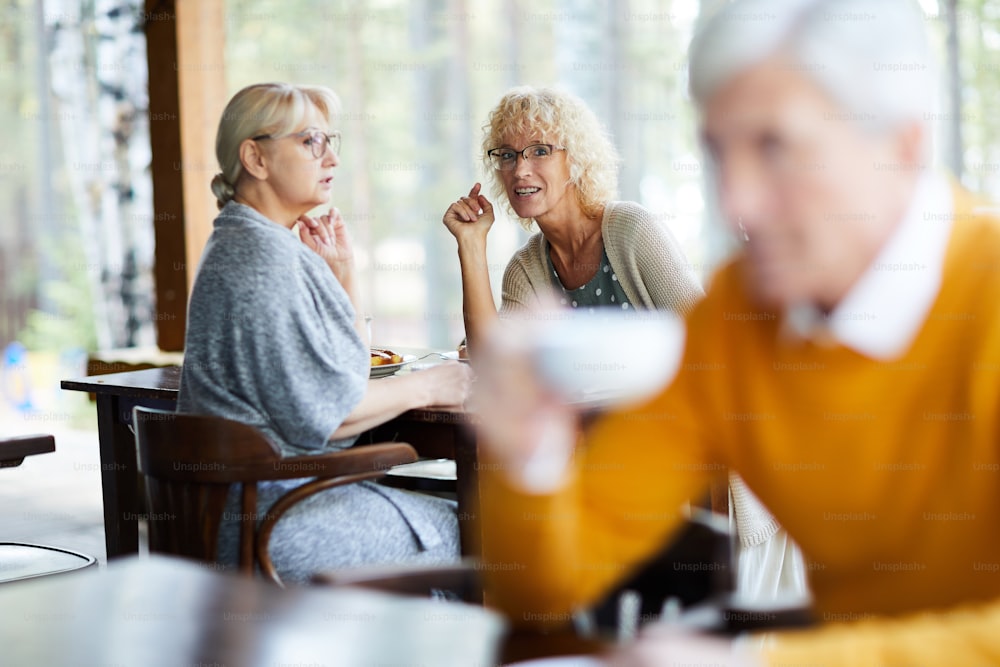 Content attractive mature ladies in casual clothing sitting at table and gossiping about handsome man in cafe, curly-haired lady pointing at this man