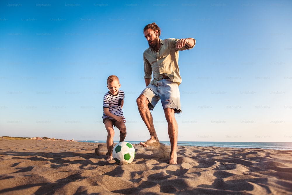 Padre e figlio felici giocano a calcio o a calcio sulla spiaggia avendo grande tempo in famiglia durante le vacanze estive.