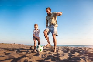 Happy father and son play soccer or football on the beach having great family time on summer holidays.