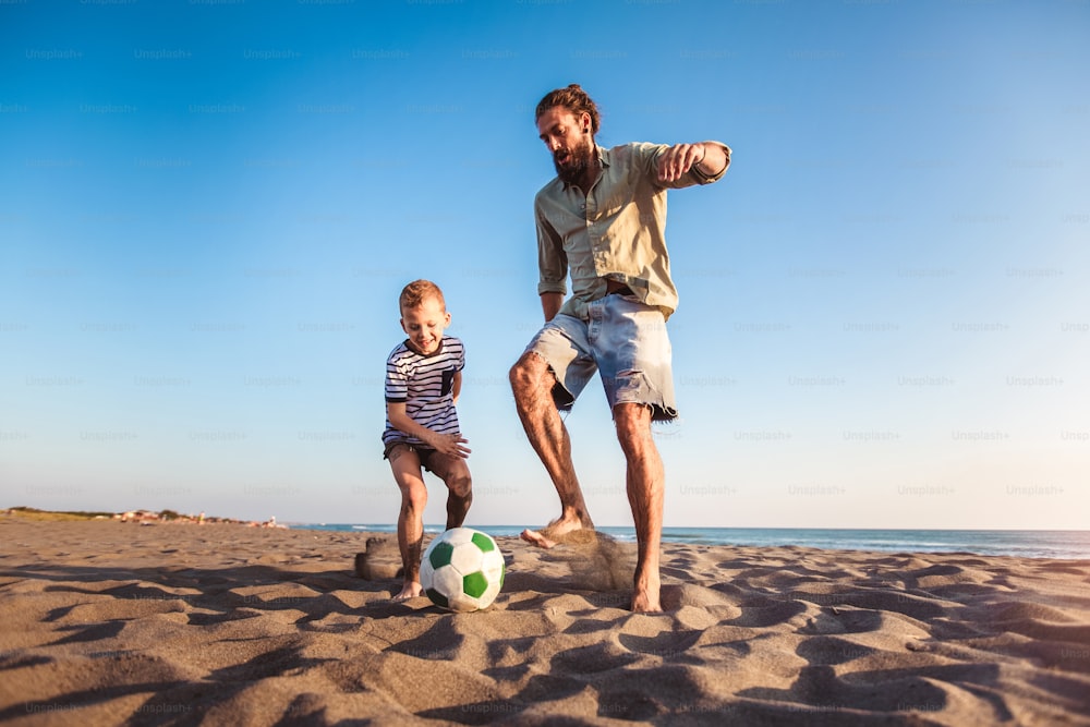 Happy father and son play soccer or football on the beach having great family time on summer holidays.