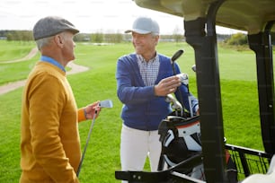 Two cheerful aged buddies in pullovers and caps discussing the forthcoming game of golf while choosing clubs
