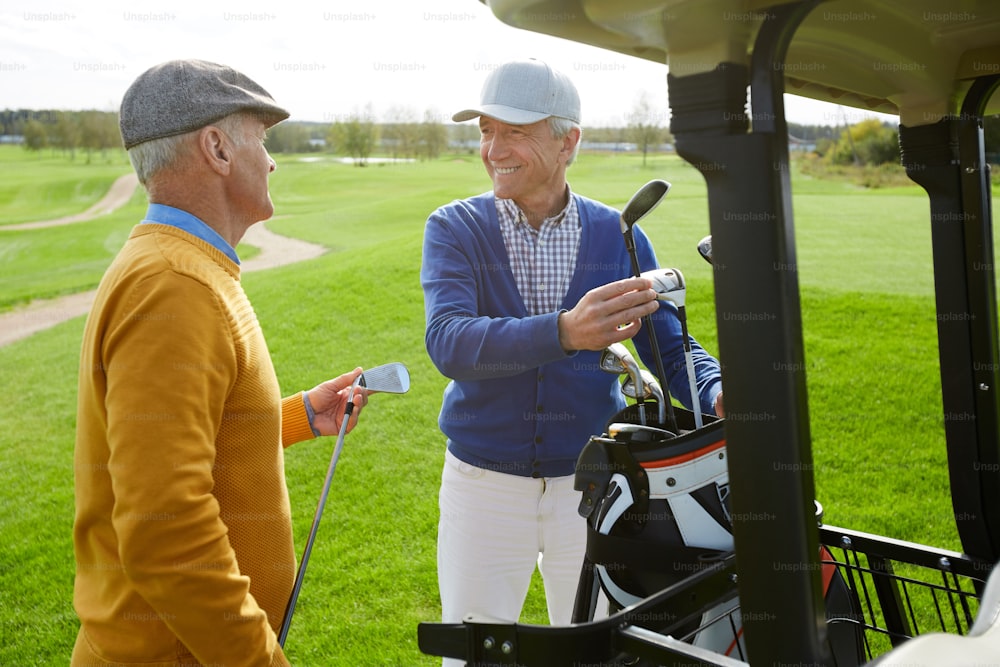 Dois alegres amigos idosos em pulôveres e bonés discutindo o próximo jogo de golfe enquanto escolhem tacos