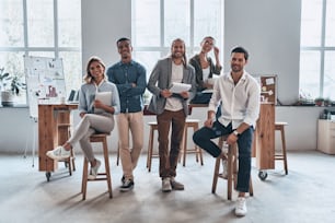 Full length of young modern people in smart casual wear smiling and looking at camera while standing in the creative office