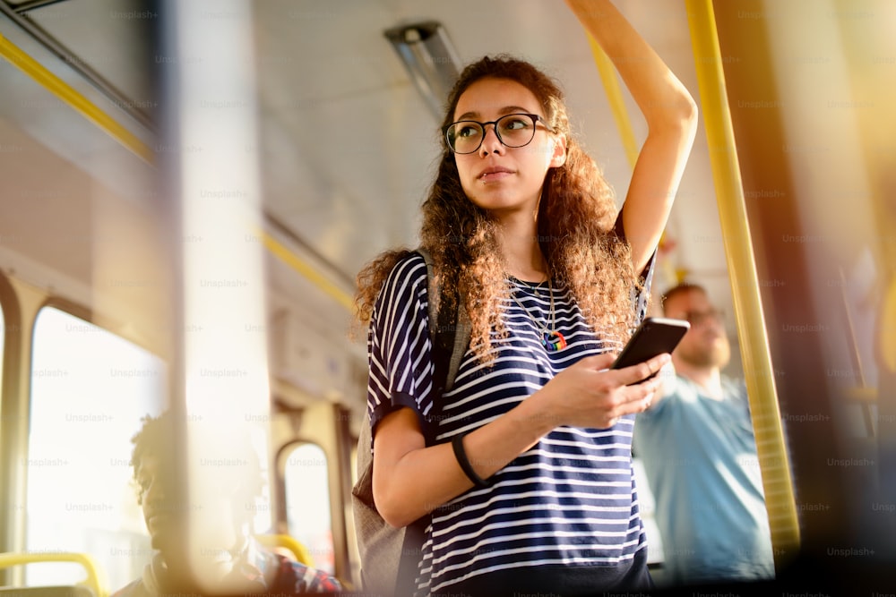 Young serious cute girl standing in a bus and holding for a bar while listening to the music.