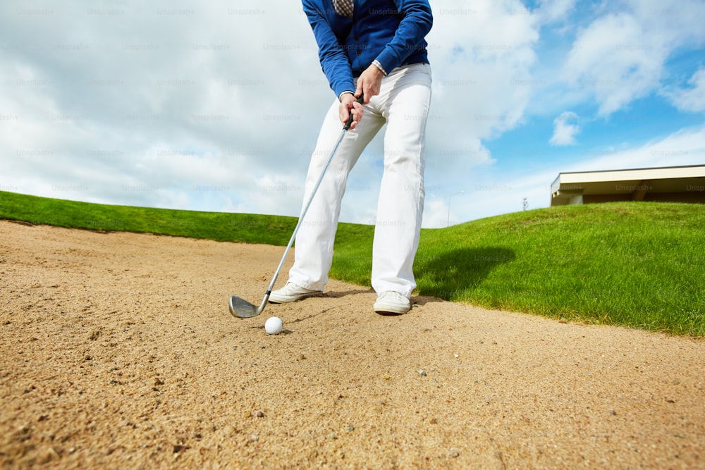 Active man in white pants and blue pullover standing on the ground and going to hit golf ball
