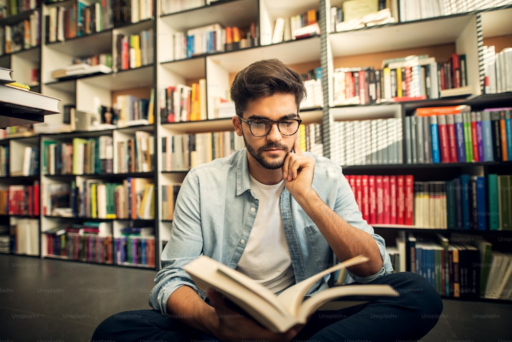 Ragazzo hipster sorridente carino seduto sul pavimento di una biblioteca e guardando attraverso alcuni libri.