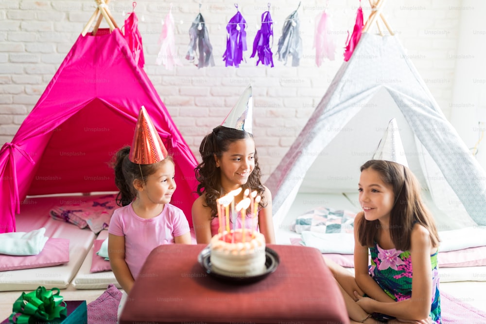 집에 있는 티피 텐트를 배경으로 테이블에 생일 케이크가 있는 파티 모자를 쓴 귀여운 소녀들