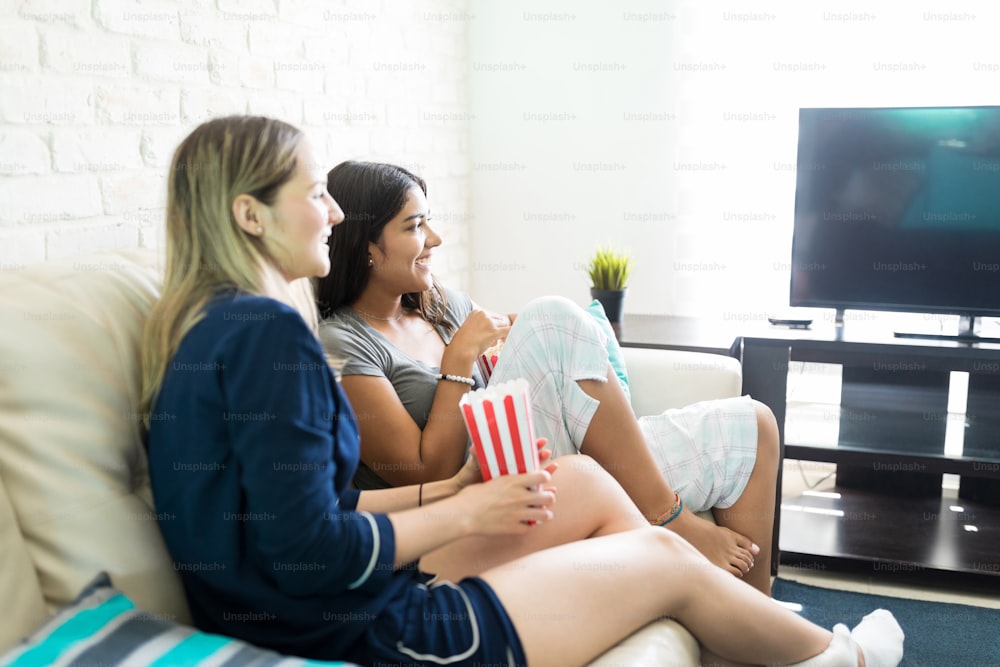 Mujeres jóvenes felices disfrutando de un programa de televisión mientras están sentadas en el sofá de su casa