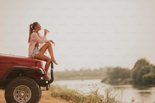 Una mujer joven se sienta en el capó de un automóvil, bebe refrescos y lo disfruta