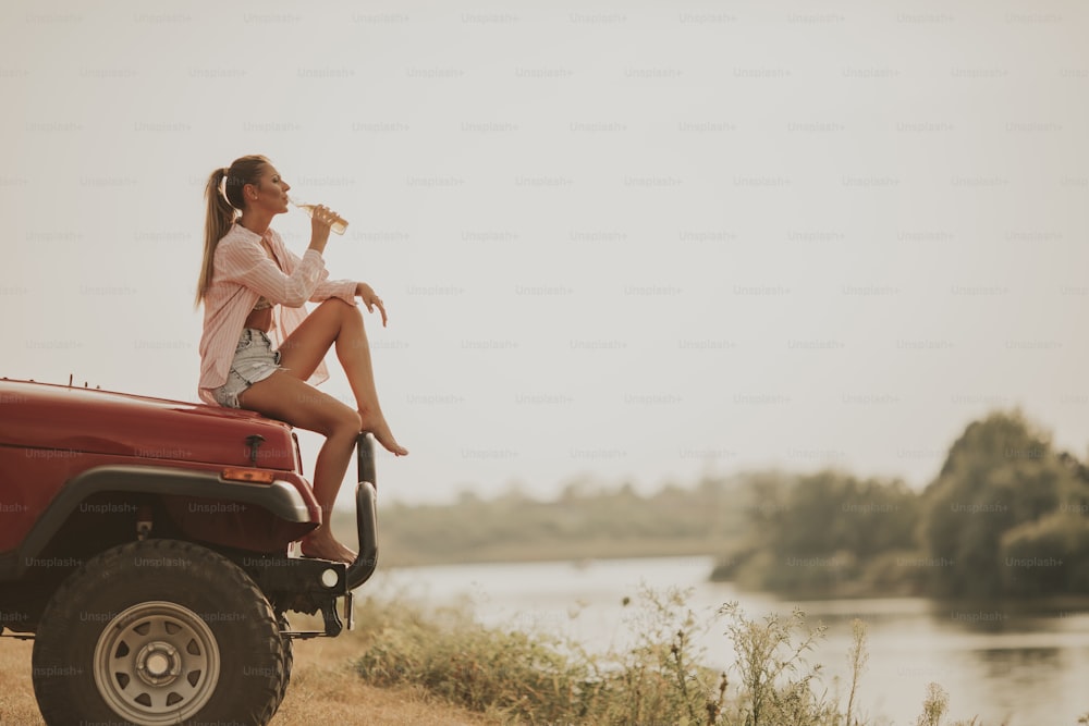 Junge Frau sitzt auf einer Motorhaube, trinkt Erfrischung und genießt es