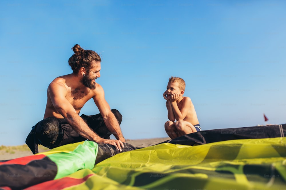 Homem surfista com seu filho em trajes de mergulho com equipamentos de kite para surfar.