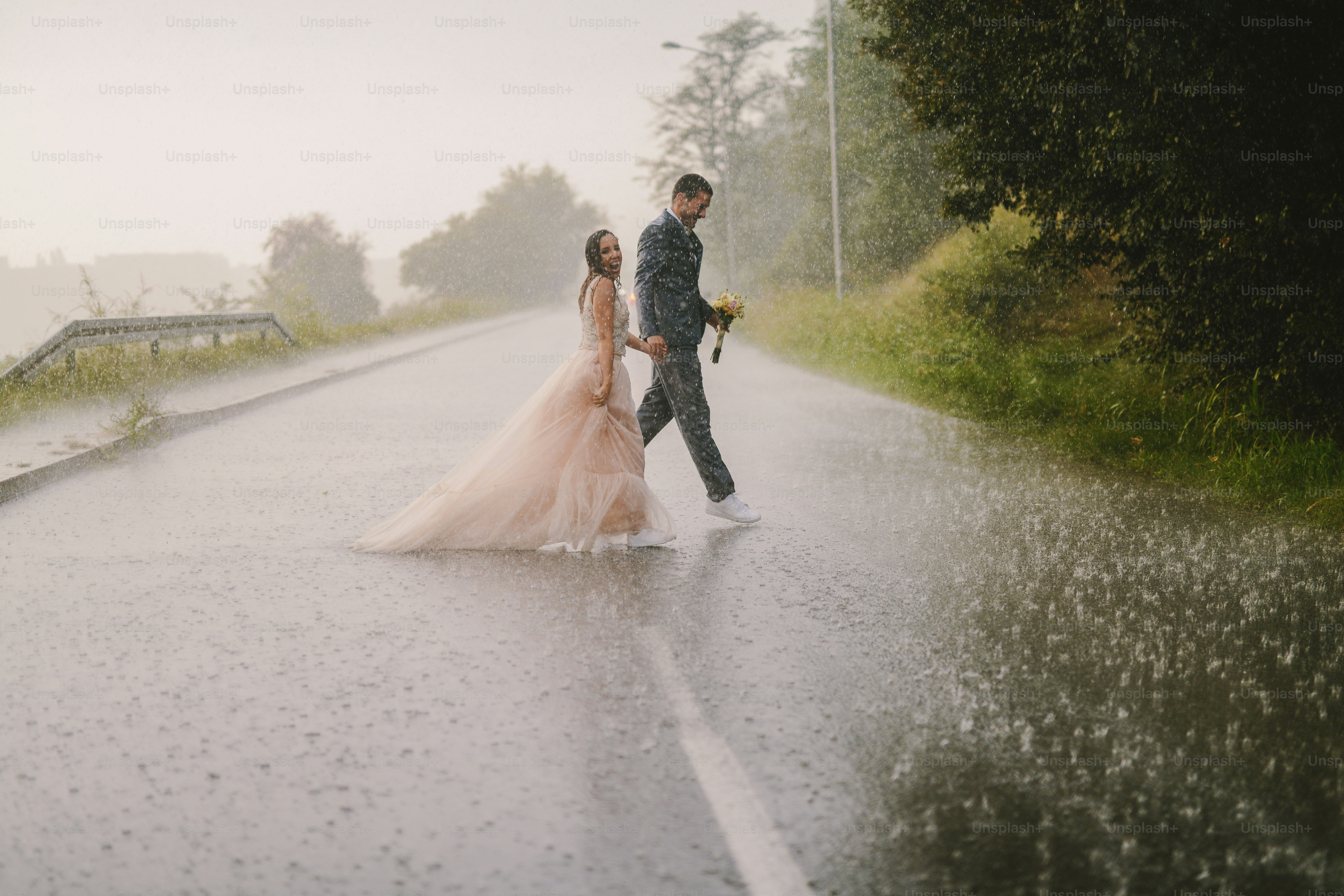 Silly young just married couple crossing road on rainy day. Walking in wet ceremonial clothes.
