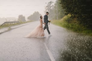 Silly young just married couple crossing road on rainy day. Walking in wet ceremonial clothes.