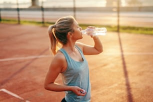 Sporty Caucasian blonde woman standing on the court and drinking water after morning exercises.