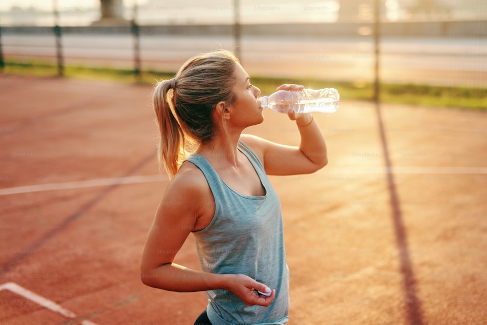 Mujer rubia caucásica deportiva de pie en la cancha y bebiendo agua después de los ejercicios matutinos.