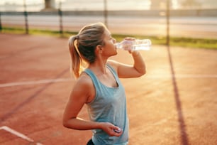 Femme blonde caucasienne sportive debout sur le terrain et buvant de l’eau après les exercices du matin.