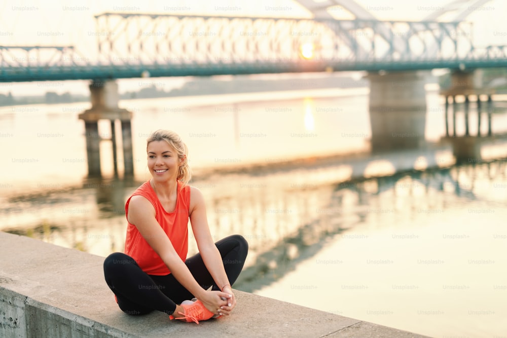 Pretty smiling blonde Caucasian woman with ponytail and dressed in sportswear sitting on the wall with crossed legs. In background bridge.