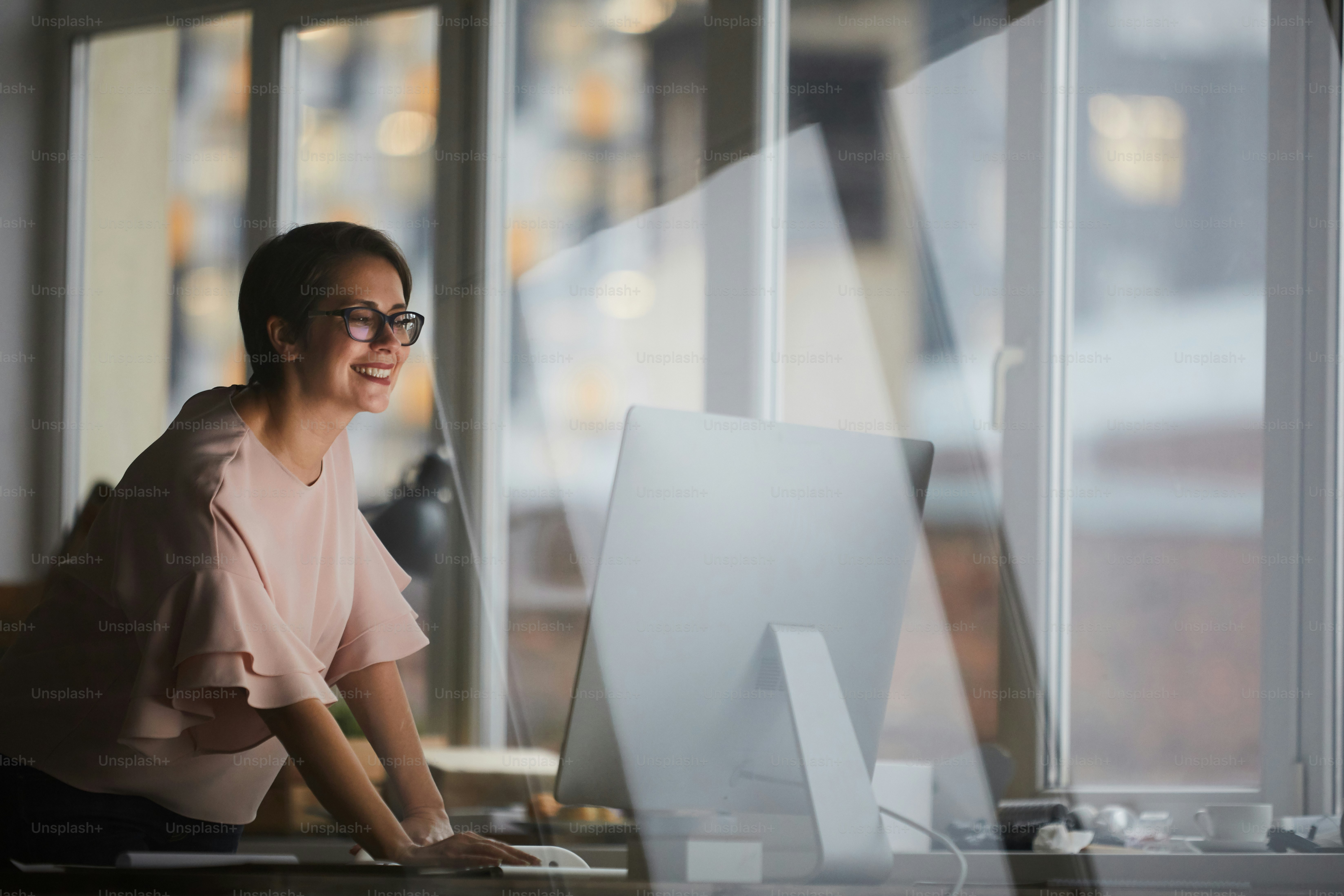 Young cheerful businesswoman leaning over table while looking at computer screen in office