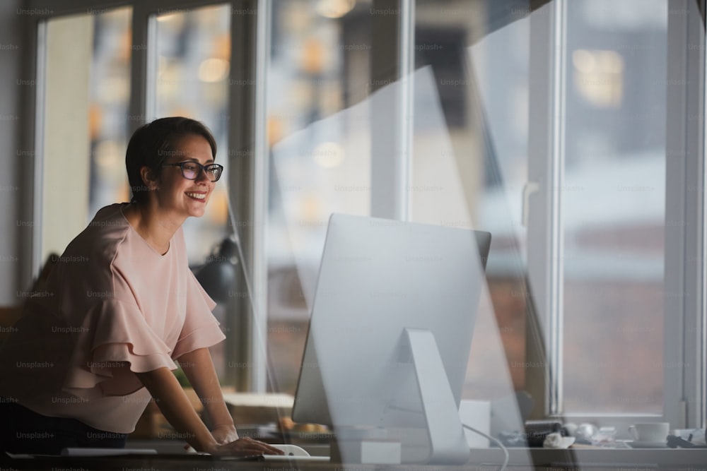 Young cheerful businesswoman leaning over table while looking at computer screen in office