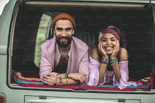 Concept of hippie lifestyle and travel. Portrait of cheerfully smiling afro-american lady and hipster male resting in back trunk of retro van