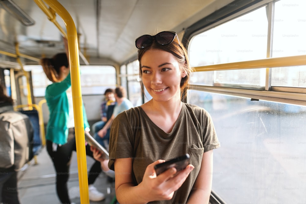 Bella ragazza caucasica sorridente con capelli castani e occhiali da sole in testa in piedi in autobus urbano e usando lo smartphone.