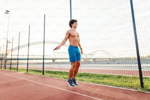 Sporty Caucasian smiling shirtless man skipping rope in court in the morning next to wire fence.