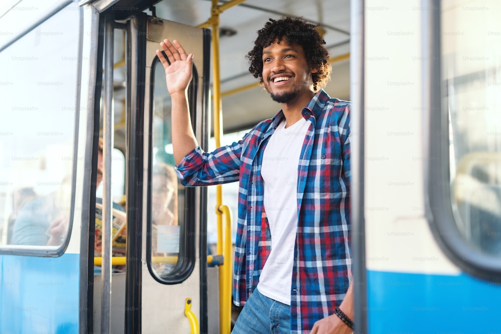 Young smiling African guy waving at girlfriend while standing on the bus door.
