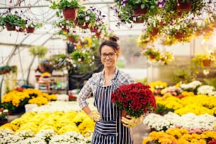 Portrait of beautiful Caucasian smiling florist holding pot with flowers while standing in greenhouse.