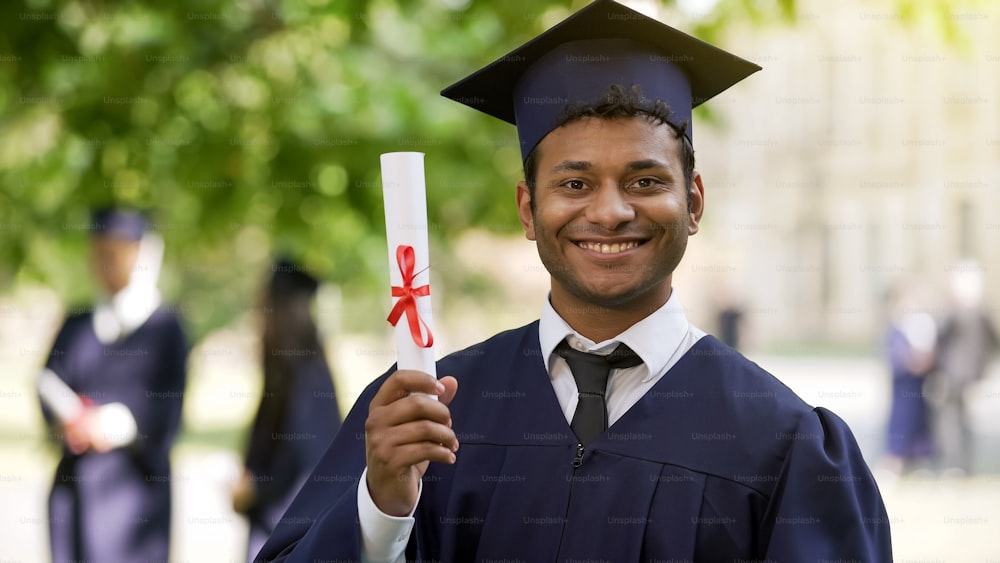 Graduado en vestimenta académica y gorra mostrando diploma y sonriendo, educación