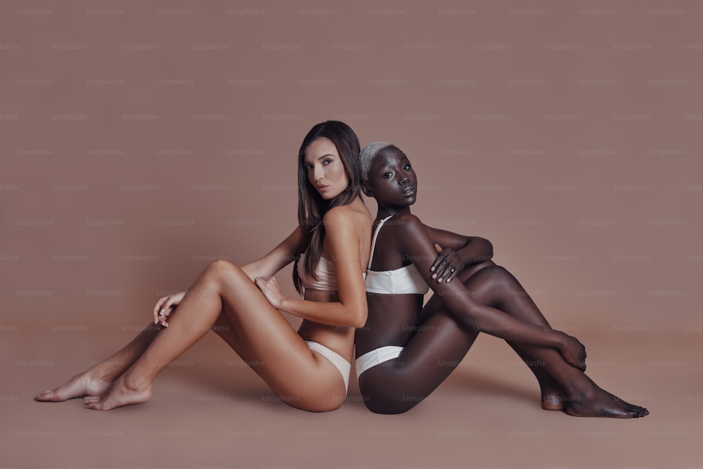Zwei attraktive gemischtrassige Frauen, die in die Kamera schauen, während sie Rücken an Rücken vor braunem Hintergrund sitzen