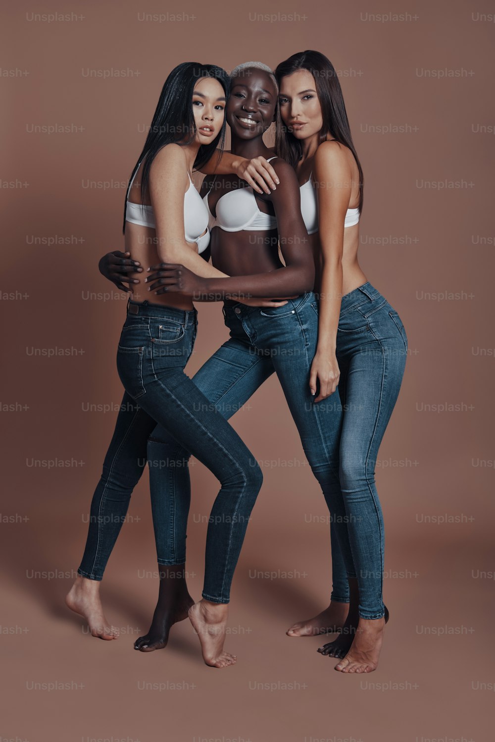 Figura intera di tre giovani donne attraenti che guardano la telecamera e sorridono mentre sono in piedi su sfondo marrone