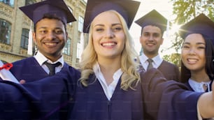 Compañeros de clase felices con vestimenta académica tomándose selfie el día de la graduación, logro