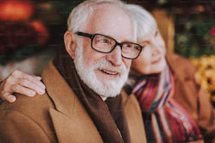 Close up portrait of stylish gentleman looking away and smiling while lady hugging him. Focus on male pensioner
